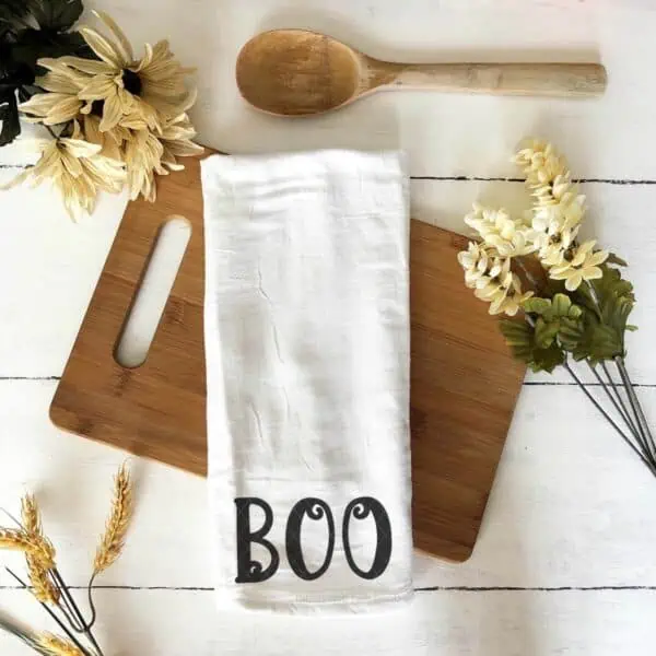 Square image of white tea towel on fall background which reads boo in black vinyl