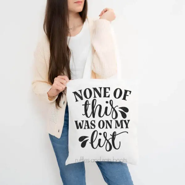 Vertical image of woman in jeans and cardigan holding a white tote bag which reads none of this was on my list.