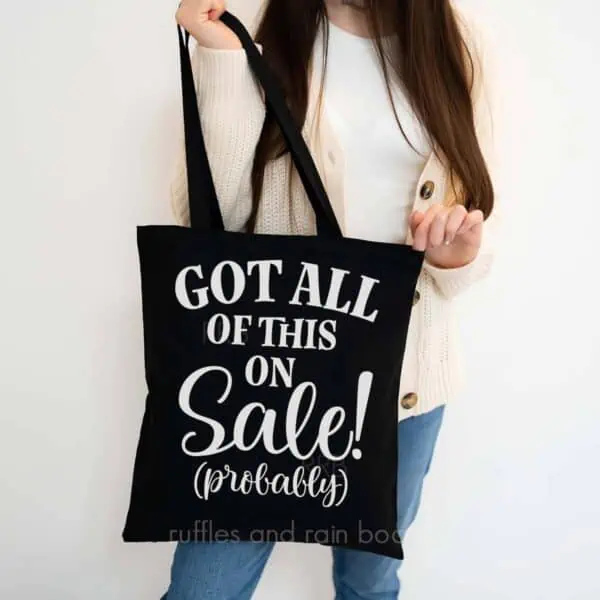 Vertical image of woman in sweater holding black tote bag which reads got all of this on sale (probably).