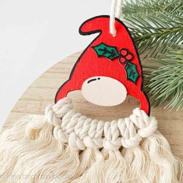 Close up, square image of a wood and macrame cord gnome ornament with red hat and holly accent.