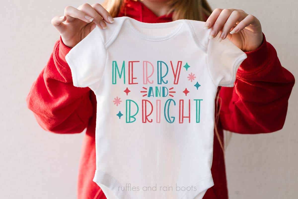 Horizontal image of woman in red sweatshirt holding a white baby body suit which reads merry and bright in colorful hues with star accents.
