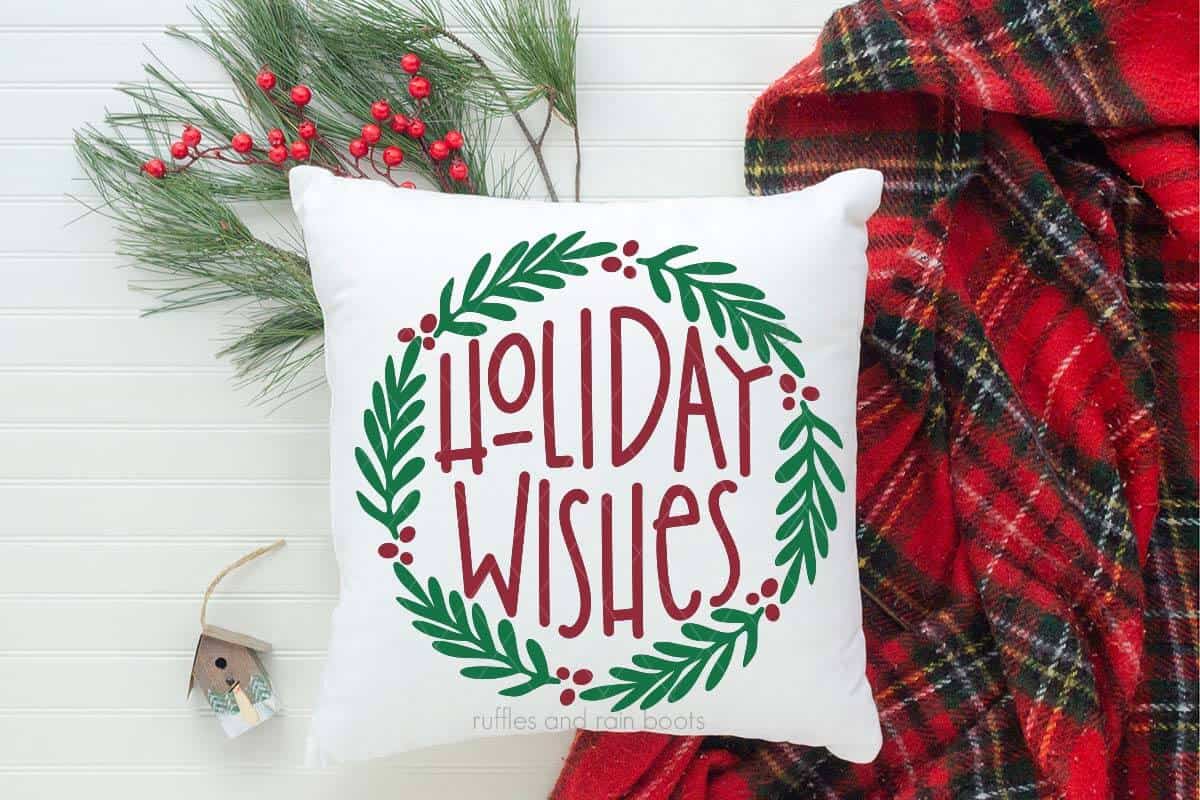 Horizontal image of holiday wishes SVG in red and green heat transfer vinyl on white pillow with plaid blanket and Christmas decorations.