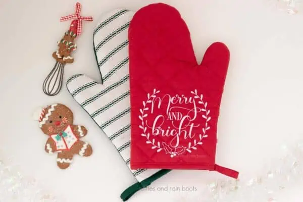 Horizontal image of red oven mitt with white heat transfer vinyl which reads merry and bright with laurel and holly.