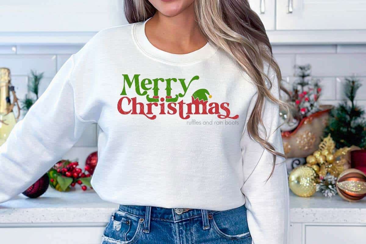 Horizontal image of woman standing in kitchen wearing jeans and a white sweatshirt which reads Merry Christmas and has elf SVG accents.