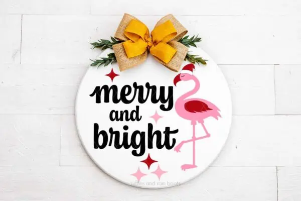 Horizontal image of white wood door sign which reads merry and bright with pink flamingo in Santa hat and a big yellow and burlap bow.