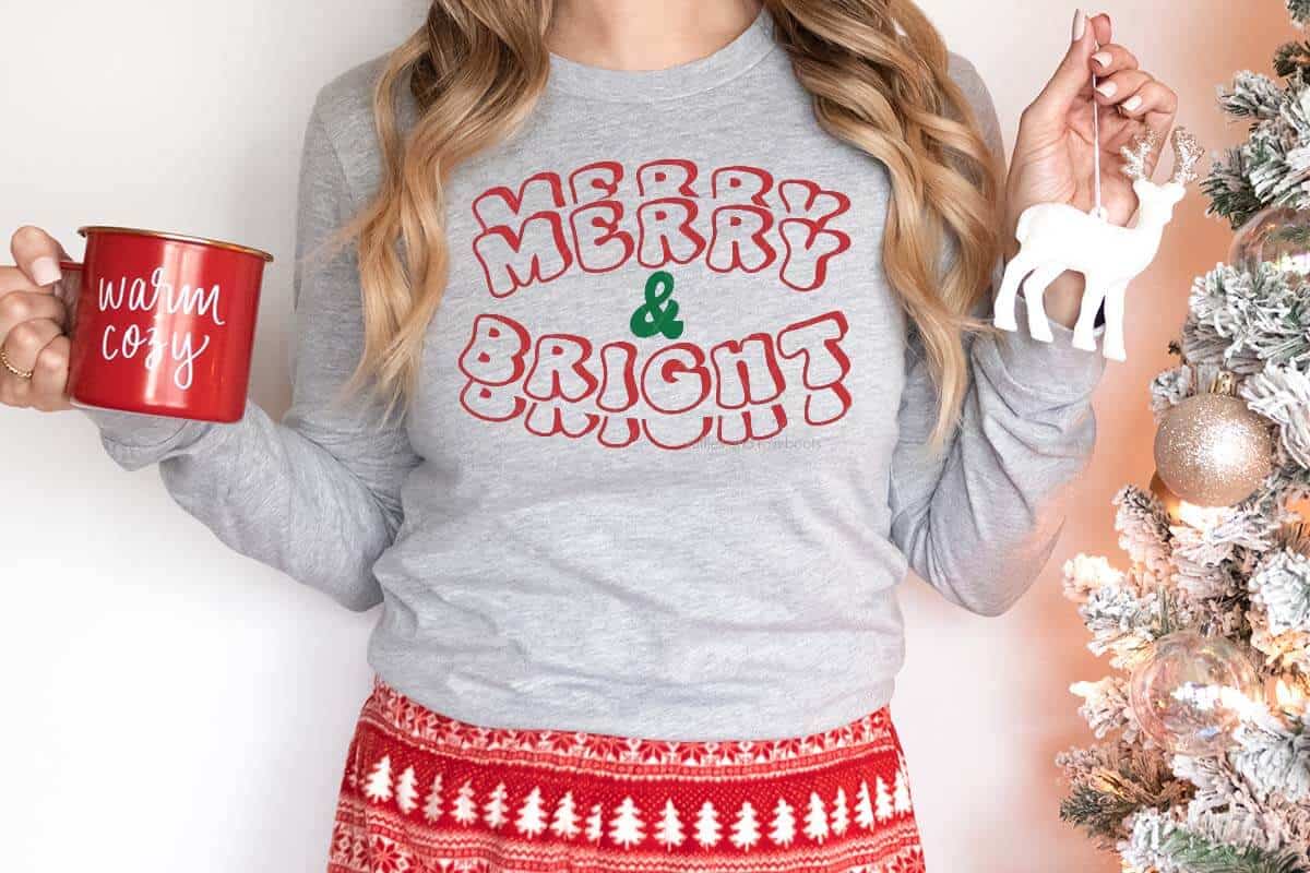 Horizontal image of woman in pajama pants and gray shirt which reads merry and bright holding a cup and ornament.