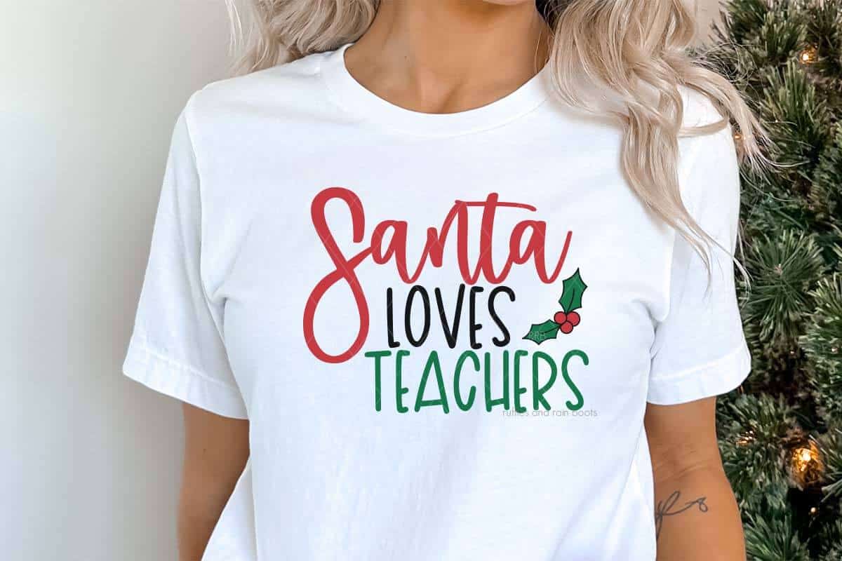 Horizontal image of woman standing next to Christmas tree wearing a white t-shirt which reads Santa Loves Teachers with holly and berries.