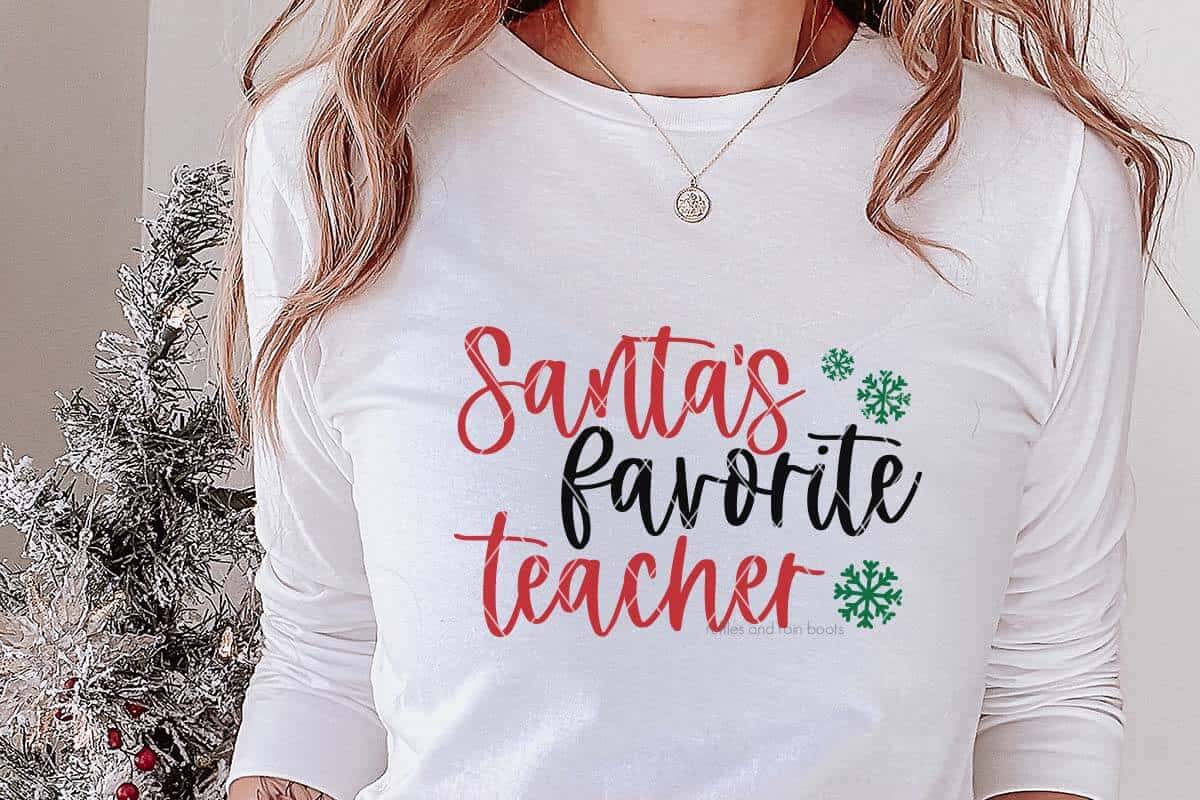 Horizontal image of woman in white t-shirt which reads Santa's favorite teacher with snowflake accent standing next to a mini Christmas tree.