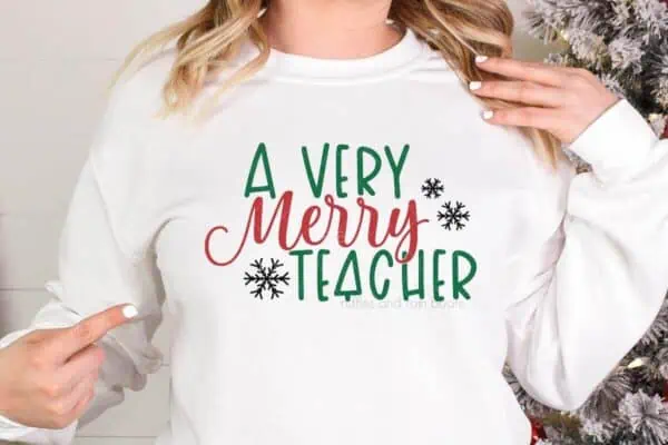 Horizontal image of woman in white sweatshirt which reads a very merry teacher standing next to a Christmas tree.