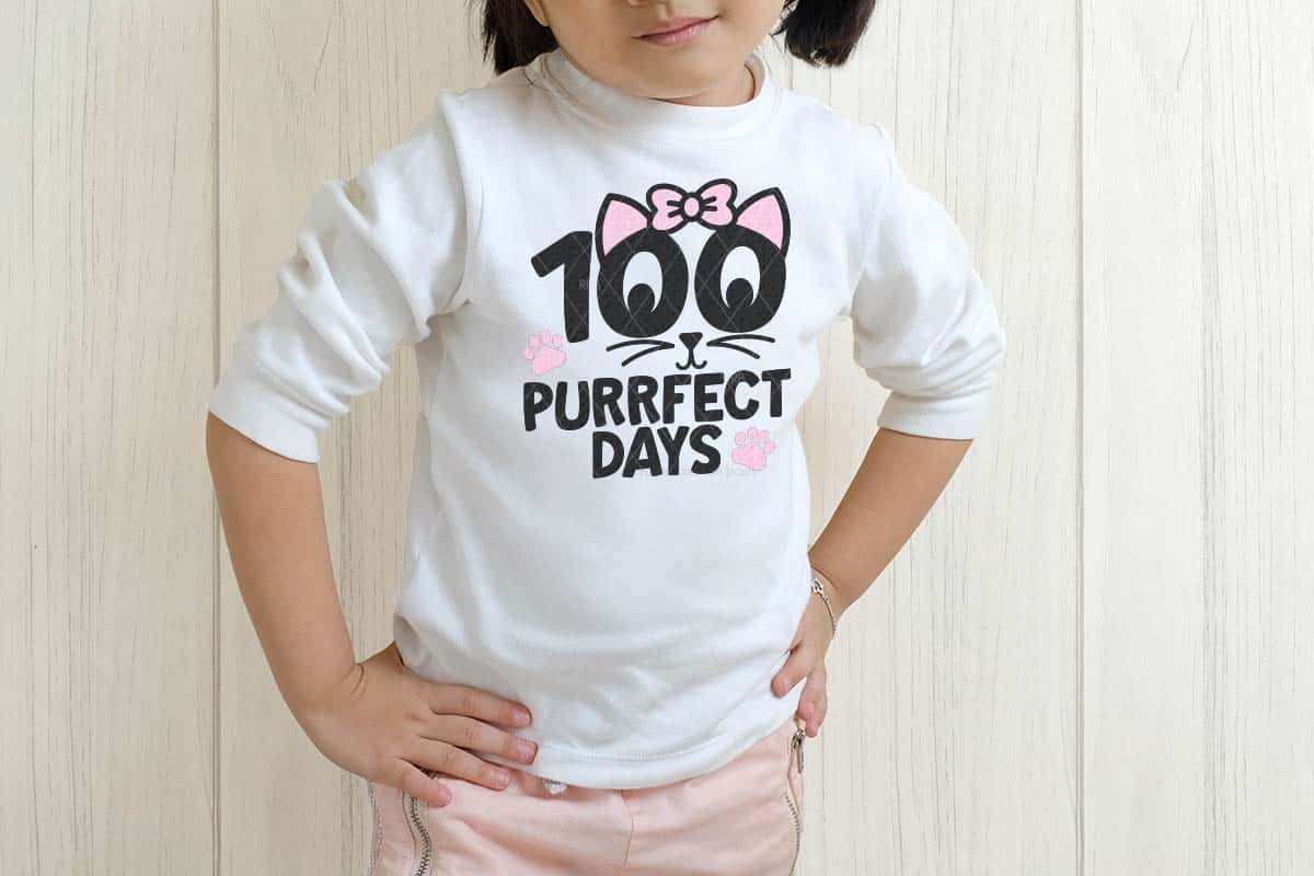 Horizontal image of a little girl standing in front of white wood panel wall wearing a white shirt which reads 100 purrfect days for the 100th day of school.