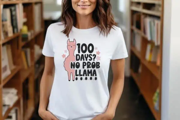 Horizontal image of woman in library wearing a white shirt which reads 100 days no prob llama featuring a 100 days of school SVG design.