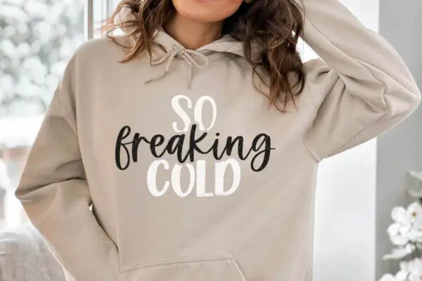 Horizontal image of woman in tan sweatshirt which reads so freaking cold standing in front of a snow covered window.