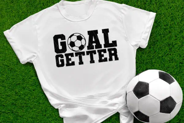 Horizontal image of a white shirt which reads Goal Getter in black vinyl on grass next to a soccer ball.