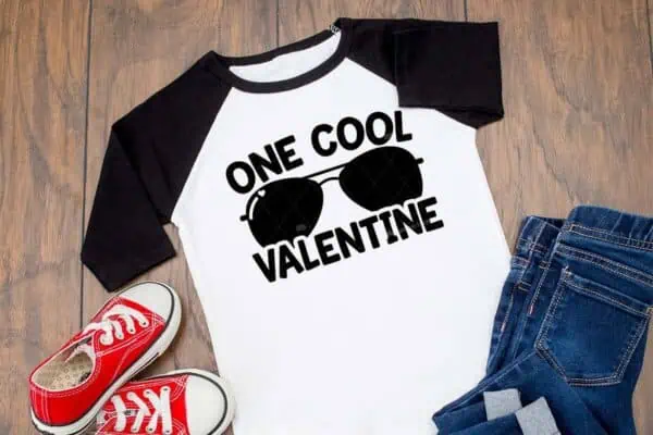 Horizontal image of wood background with black raglan t shirt which reads one cool Valentine with boys mini red tennis shoes.