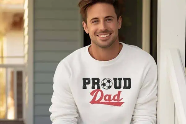 Horizontal image of a man in a white sweatshirt which reads proud soccer dad sitting on a front porch.