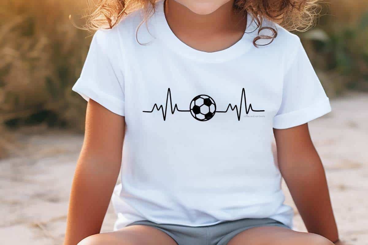 Horizontal image of a child outside wearing a white shirt with a soccer heartbeat SVG done in black HTV with a Cricut.