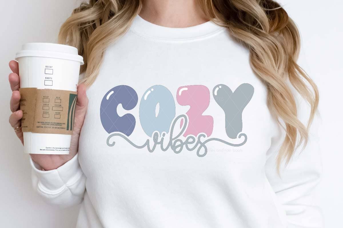 Horizontal image of woman holding coffee cup wearing a white sweatshirt which reads cozy vibes in pastel wintery colors of heat transfer vinyl