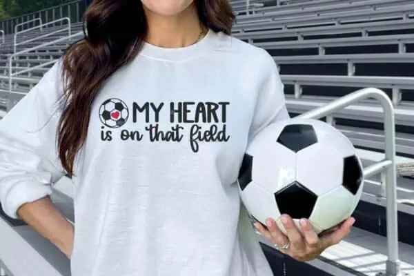 Horizontal image of a woman in a white sweatshirt which reads my heart is on that field with soccer ball SVG standing in front of stands holding a soccer ball.