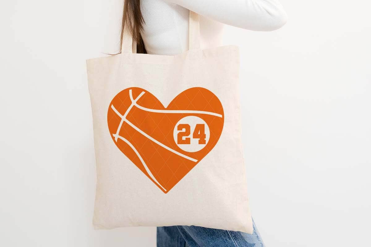 Horizontal image of a woman in jeans and a white t-shirt holding a tote bag with a bright orange basketball heart with the player number 24.
