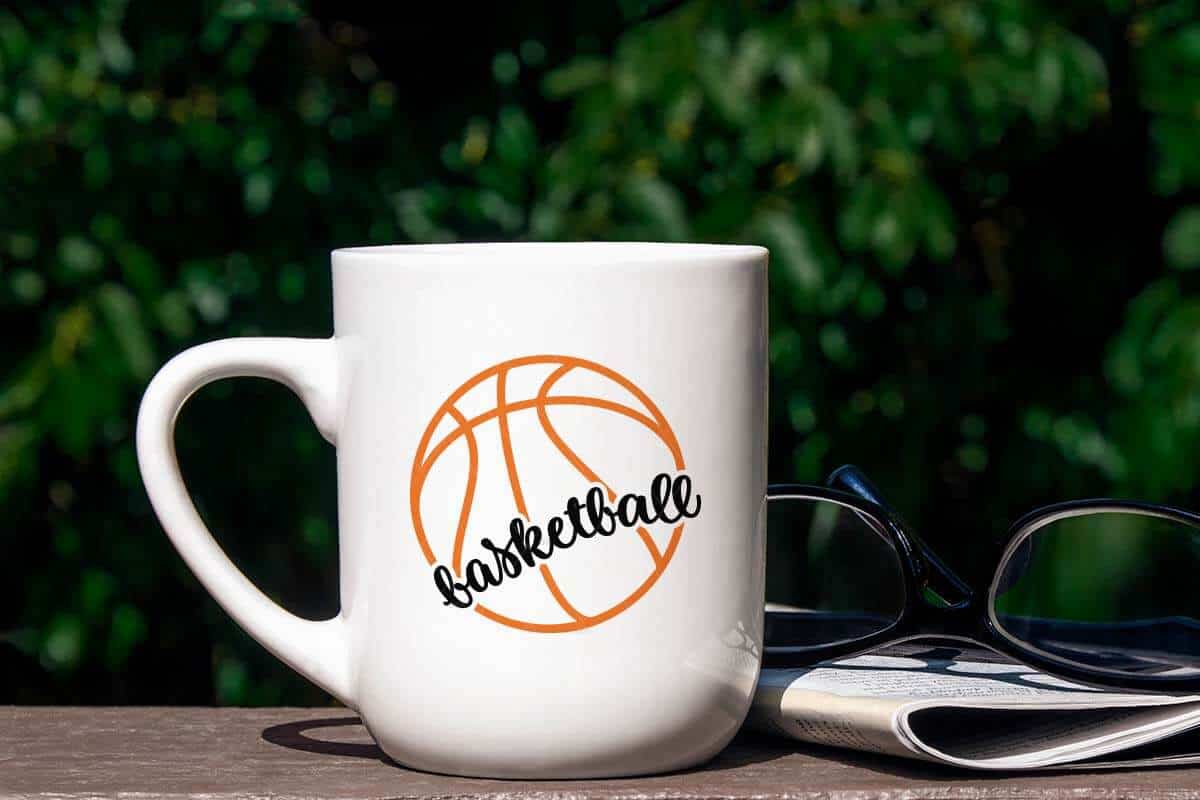 Horizontal image of a mug with a basketball outline SVG and script basketball in orange and black permanent vinyl on a porch in a sunny outdoor space next to sunglasses and a book.