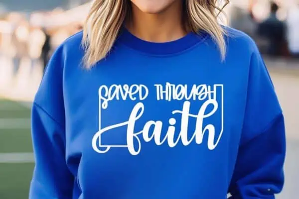 Horizontal image of woman in park wearing a blue sweatshirt which reads saved through faith in white vinyl.