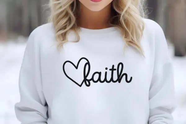 Horizontal image of woman outside wearing a white sweatshirt with faith and a heart in black vinyl.