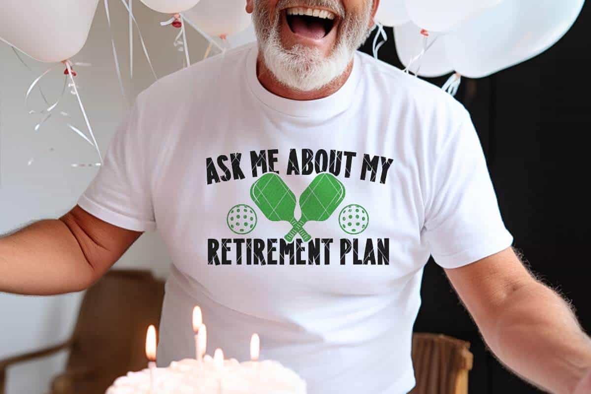 Horizontal image of an older man in front of balloons and cake with a white t-shirt which reads ask me about my retirement plan with pickleball paddles and dink balls.