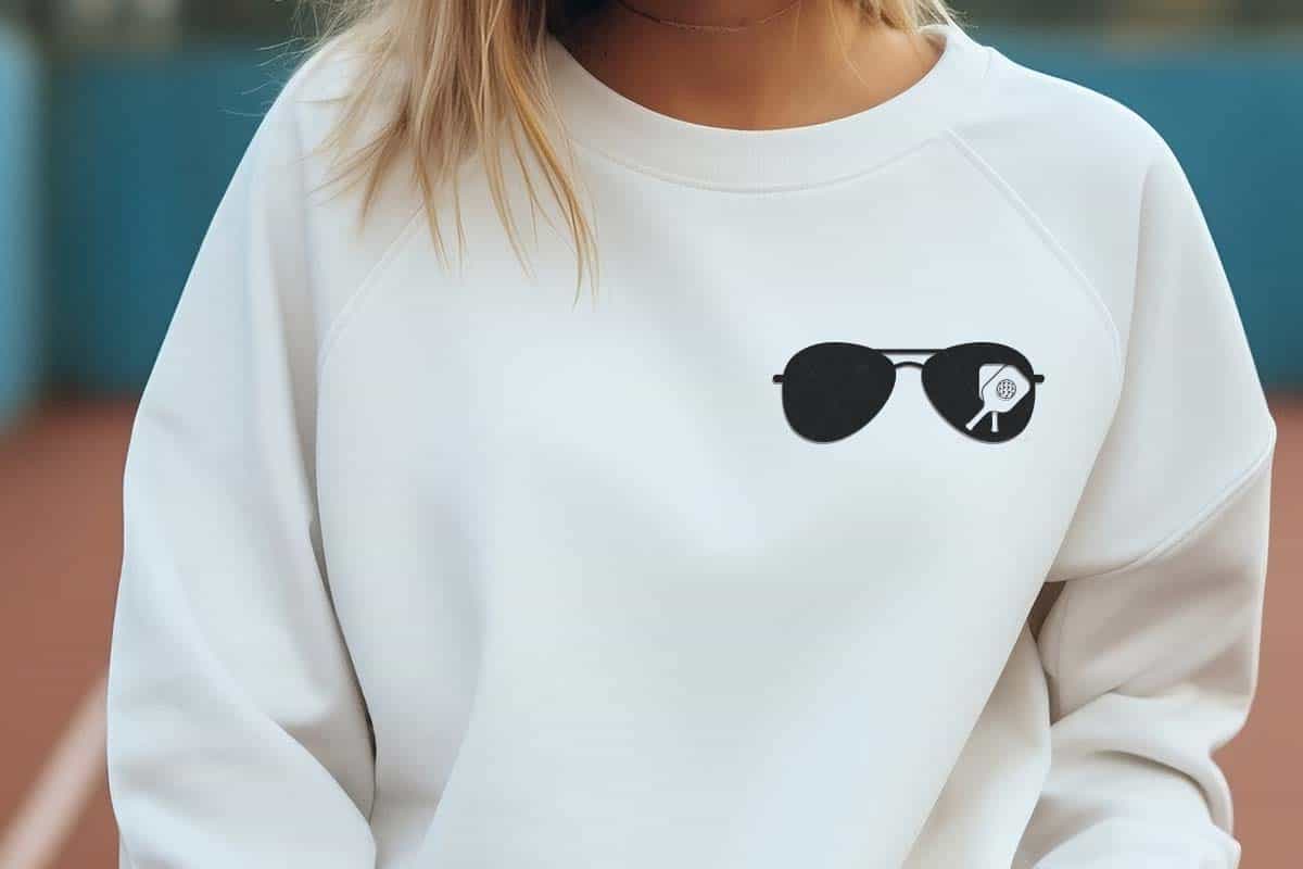 Horizontal image of a woman on a tennis court wearing a white sweatshirt with a pocket sized pickleball sunglasses design.