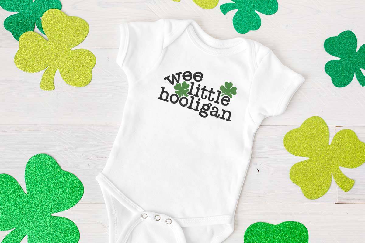 Horizontal image of a white wood background with shamrocks and a white baby body suit with the words wee little hooligan and four leaf clovers.