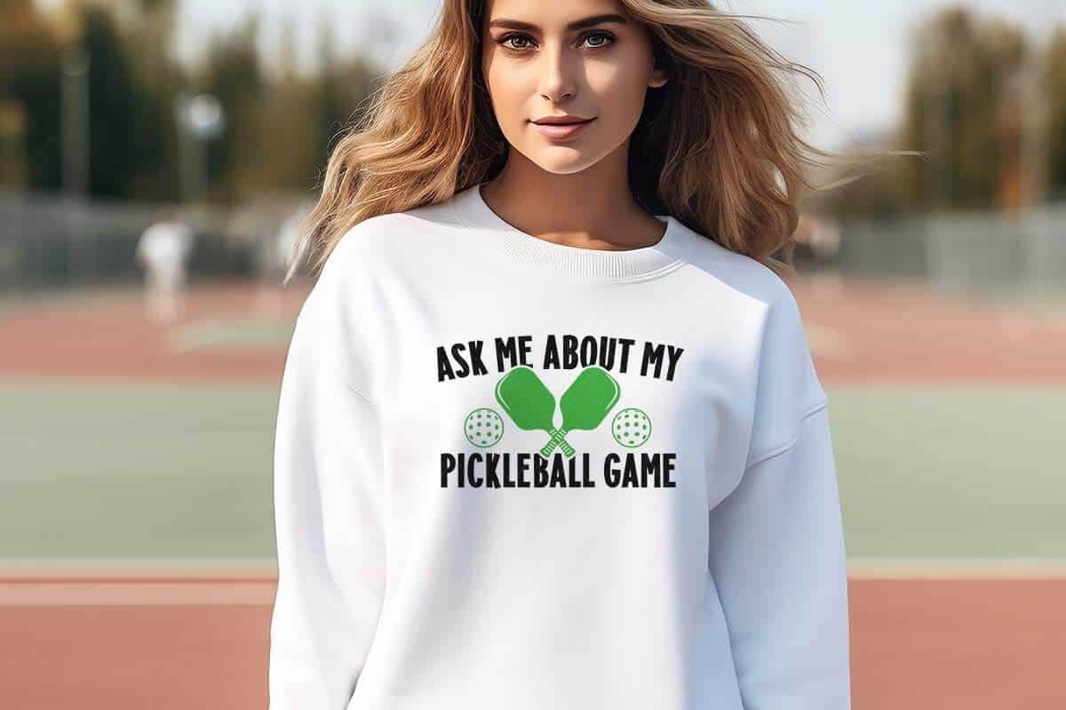 Horizontal image of a woman on a tennis court wearing a white sweatshirt which reads ask me about my pickleball game with two paddles and dink balls.