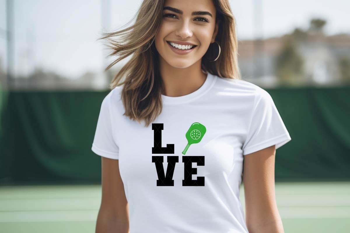 Horizontal image of a smiling woman on a pickleball court wearing a white t-shirt which reads LOVE with a pickleball paddle and dink ball in place of the O.