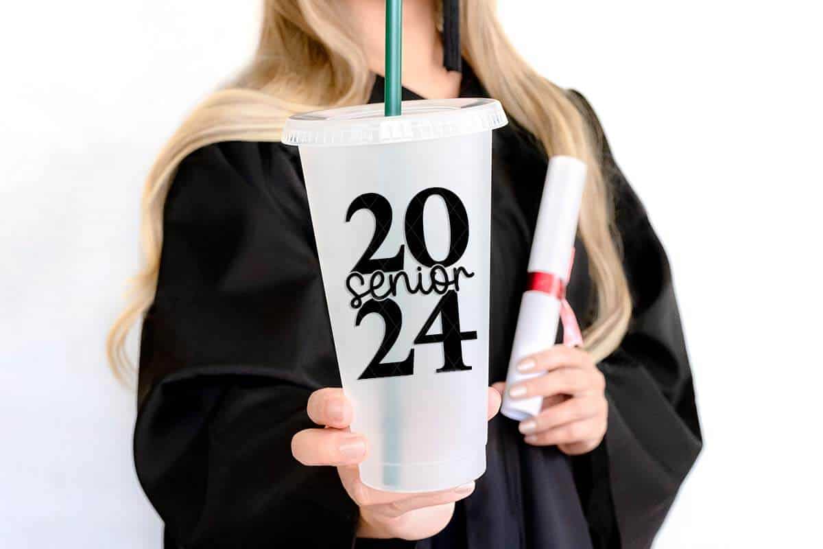 Horizontal image of a graduate holding a degree scroll and a cold cup with senior 2024 on it in black vinyl.