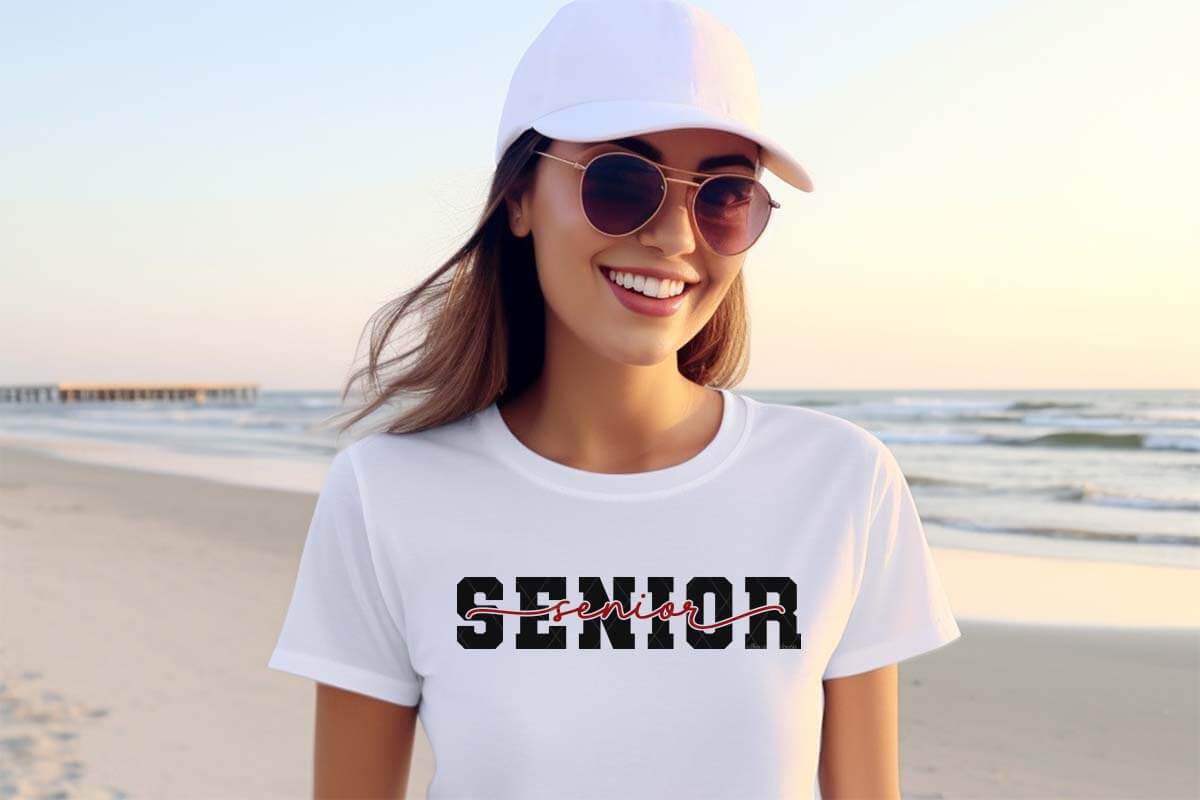 Horizontal image of a high school senior on a beach wearing sunglasses, a white hat, and a white shirt which reads senior in a varsity and offset style.