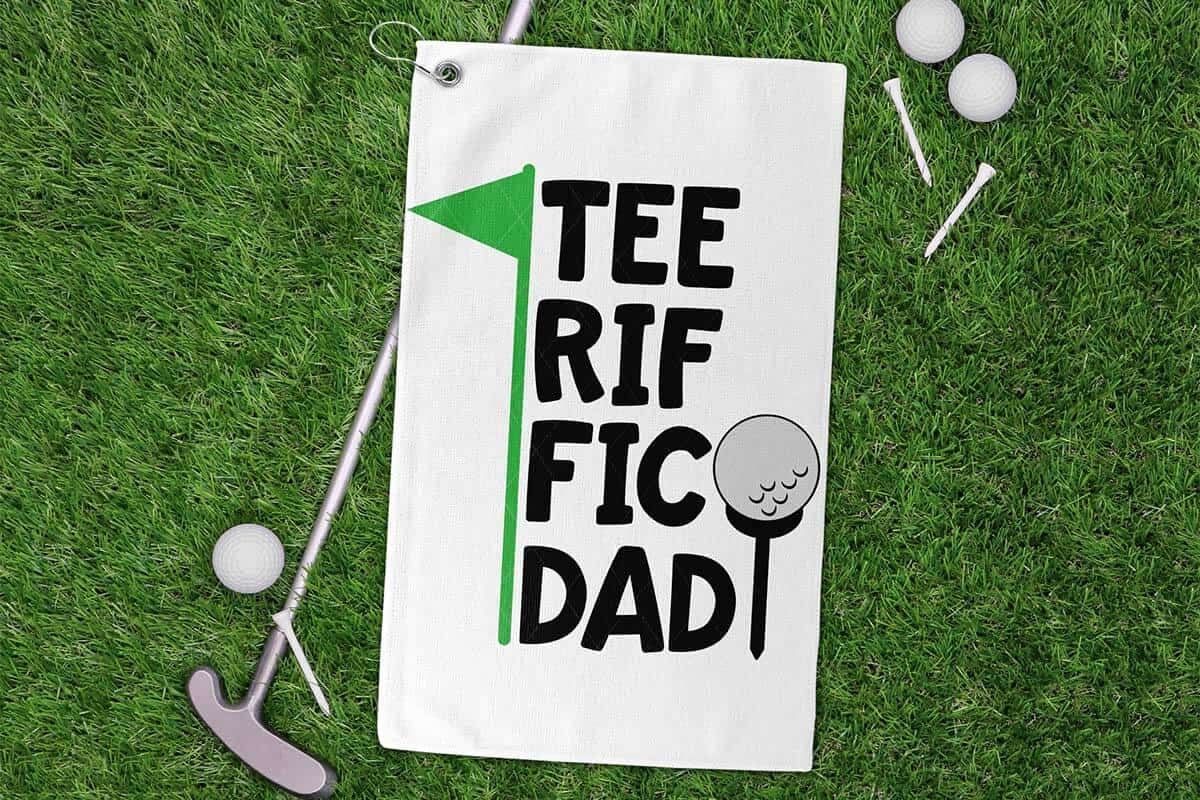Horizontal image of a golf towel on grass with a putter, golf balls, and tees which reads teeriffic Dad.