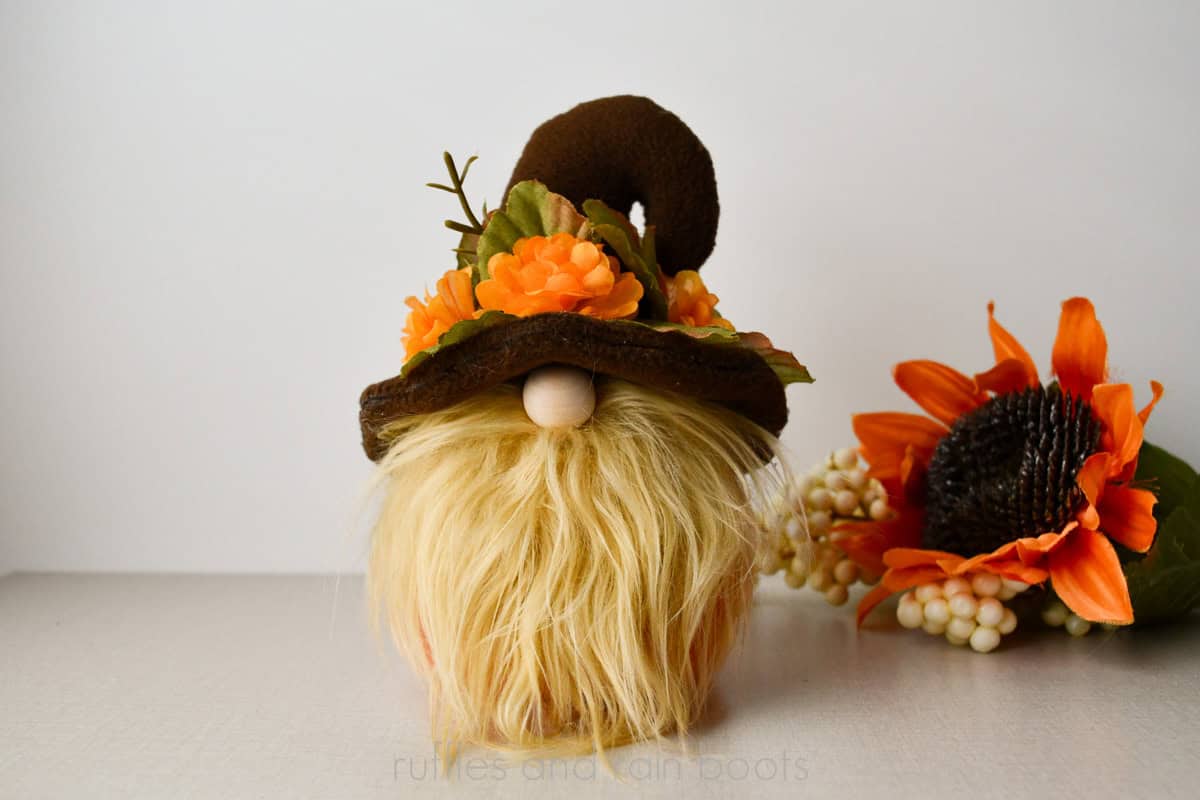 Horizontal image of a brown hook hat gnome with a brim of orange blossoms and leaves with a tan gnome beard and sunflower background.