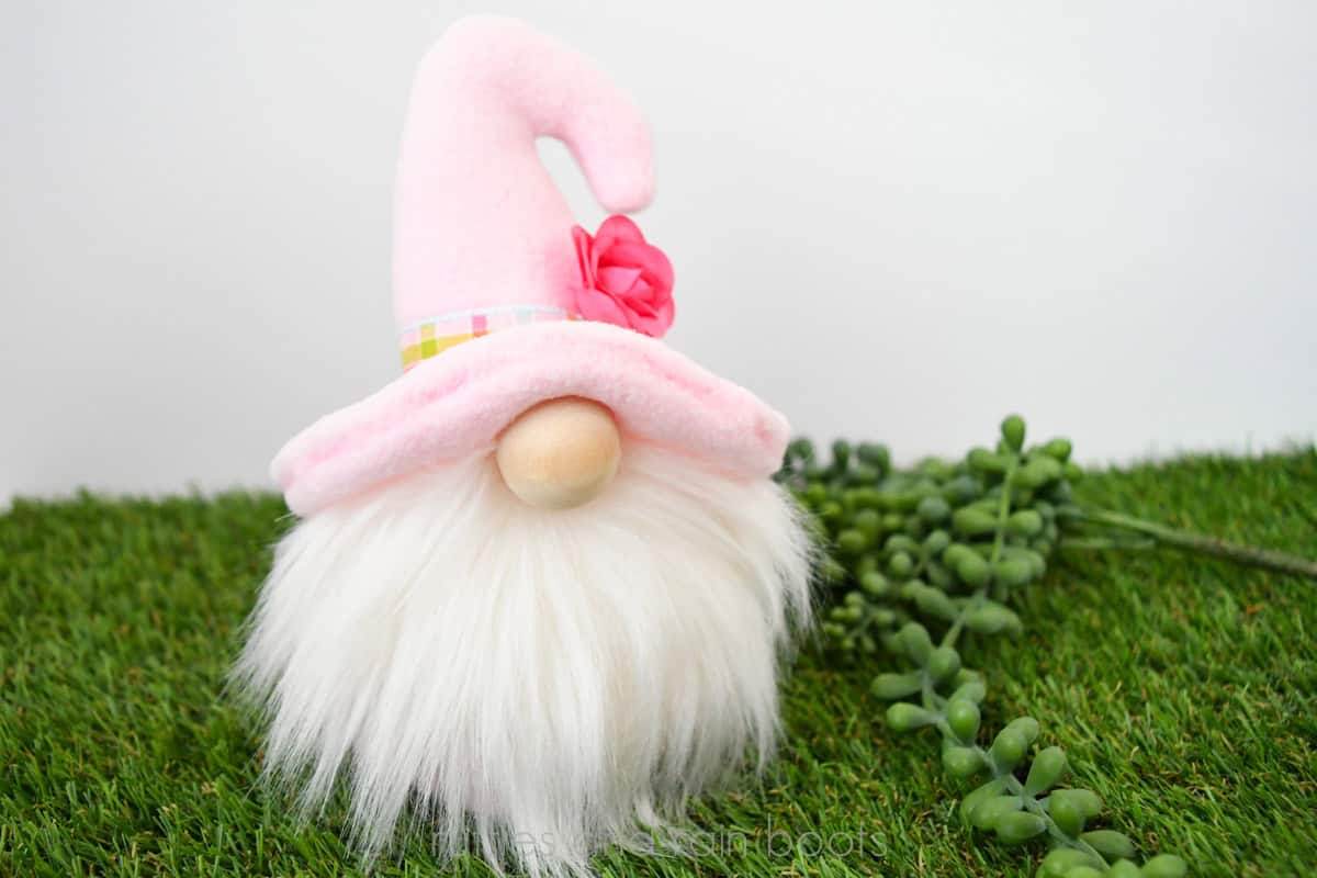Horizontal image of a spring gnome with a pink hooked hat, ribbon and flower brim, and a white gnome beard on a grass background.