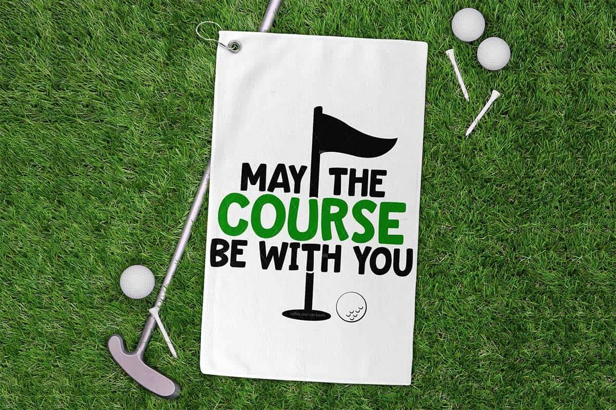 Horizontal image of a golf club, tees, and golf balls on grass with a golf towel which reads may the course be with you.