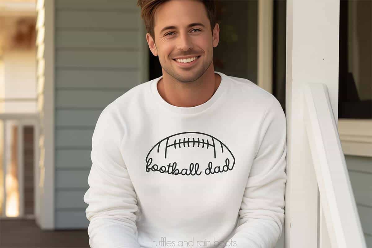 Horizontal image of man sitting on a porch wearing a white sweatshirt which reads football dad in a hand drawn simple design.