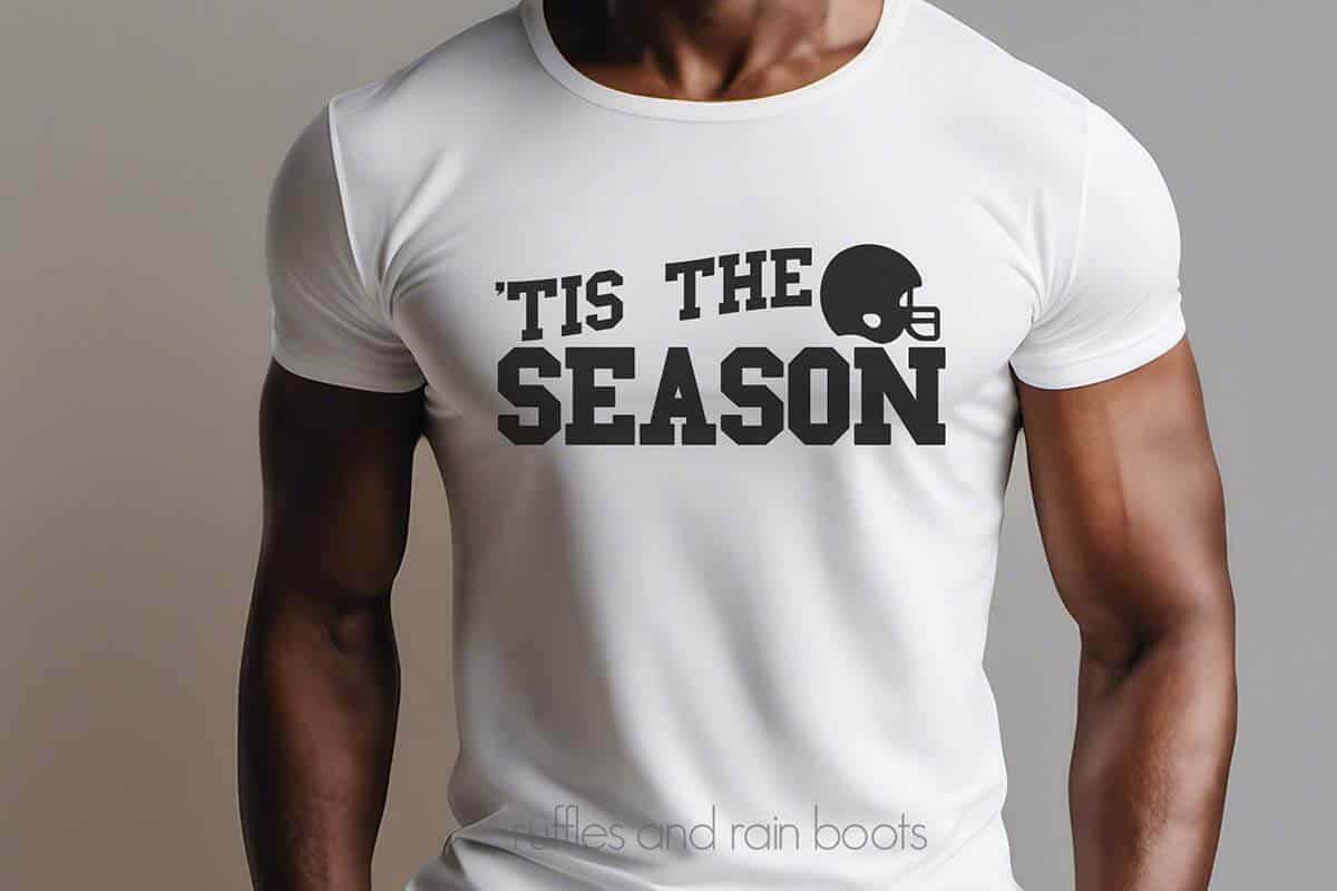 Horizontal image of a man in a white t-shirt with black lettering which reads tis the season with a football helmet.