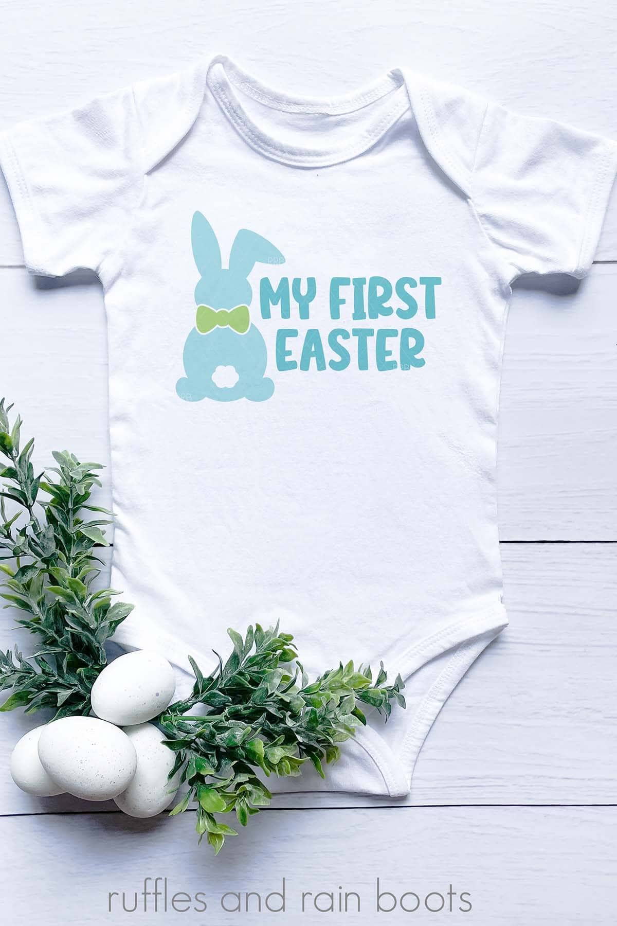 Vertical image of a white baby bodysuit with My First Easter SVG in blue with a green bow tie.