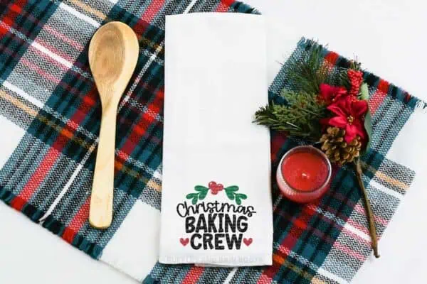 Horizontal image of a Christmas baking crew SVG design with hearts and holly frame on a white kitchen towel on a plaid background.