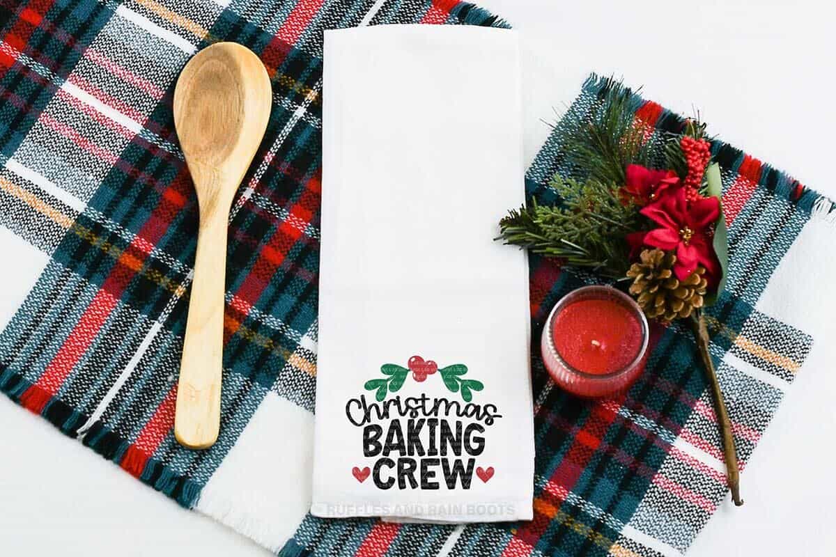 Horizontal image of a Christmas baking crew SVG design with hearts and holly frame on a white kitchen towel on a plaid background.