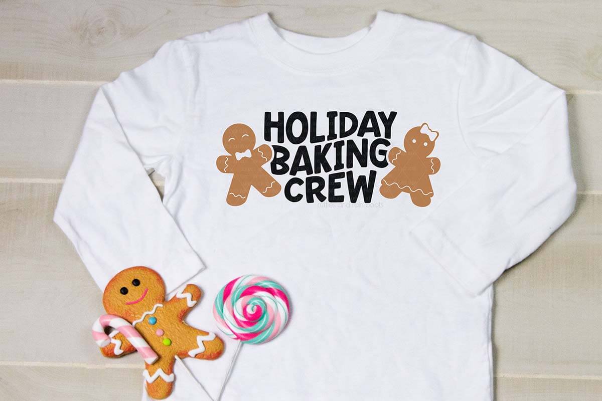 Horizontal close up image of a child's white t-shirt with holiday baking crew in black vinyl and two gingerbread cookie SVG designs on either side.