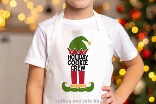 Horizontal image of a little boy in front of a Christmas tree wearing a white apron with a little elf and a design which reads holiday cookie crew.