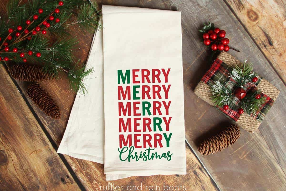 Horizontal image of a Christmas wooden table with a kitchen towel which reads Merry Merry Merry Merry Merry Christmas in red and green heat transfer vinyl.
