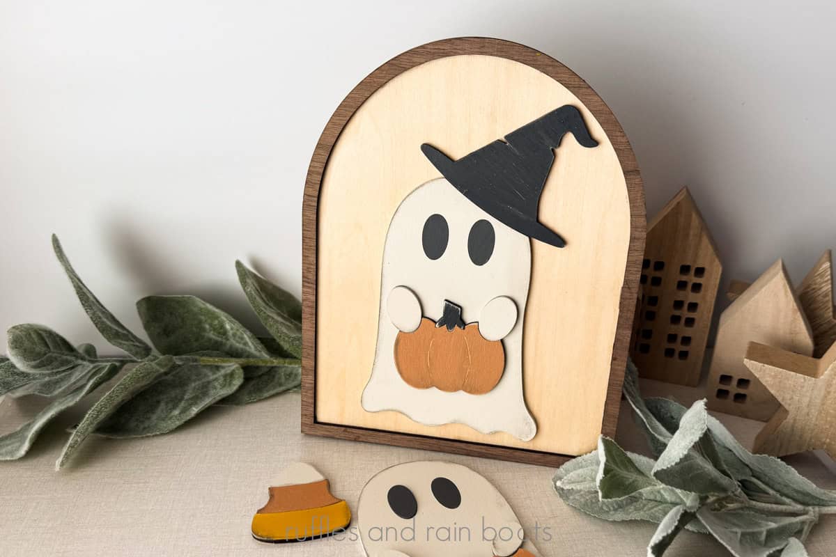 Horizontal image of a ghost wearing a witch hat, holding a pumpkin in a framed door sign made on a diode laser using plywood.