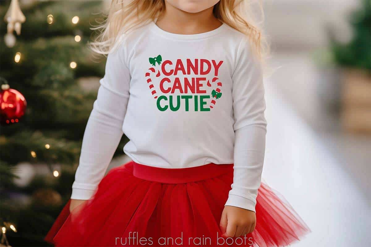 Horizontal image of a little girl wearing a white long sleeve shirt with candy cane cutie in red, white, and green vinyl standing in front of a decorated Christmas tree.